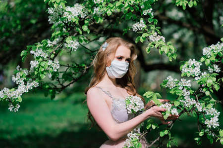 Beautiful young bride in a wedding dress and a white medical mask on her face near a blooming magnolia. Covid-19 protects.の写真素材
