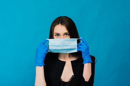Cheerful and joyful young female playing with protective medical mask and gloves on blue background....