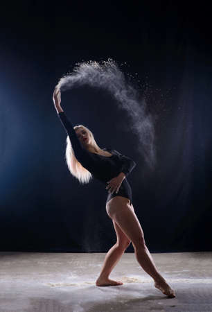 Beautiful plump blonde girl wearing a black gymnastic bodysuit covered with clouds of the flying white powder jumps dancing sitting on a dark. Artistic conceptual and advertising photo. Copy spaceの写真素材