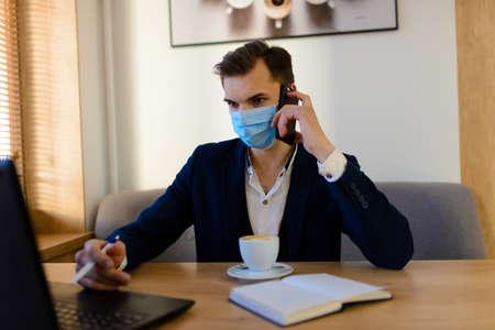 Portrait of young handsome businessman with face mask in quarantine of flu. Photo in caffee with laptop, mobile phone. Coronavirus pandemic.の写真素材