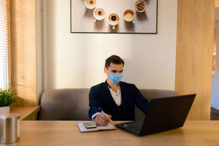 Portrait of young handsome businessman with face mask in quarantine of flu. Photo in caffee with laptop, mobile phone. Coronavirus pandemic.の写真素材