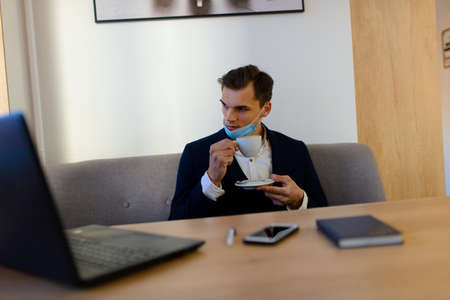 Portrait of young handsome businessman with face mask in quarantine of flu. Photo in caffee with laptop, mobile phone. Coronavirus pandemic.の写真素材