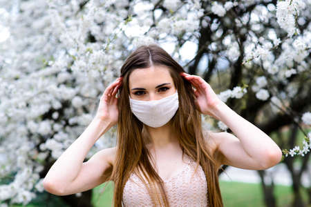 Close up portrait of tender girl in a white blouse under a blossoming cherry tree with a mask with flowers on from the coronavirus.の写真素材
