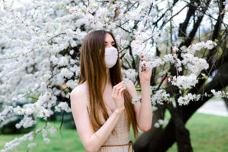 Close up portrait of tender girl in a white blouse under a blossoming cherry tree with a mask with flowers on from the coronavirus.の写真素材
