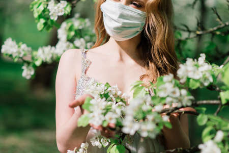 Beautiful young bride in a wedding dress and a white medical mask on her face near a blooming magnolia. Covid-19 protects.の写真素材