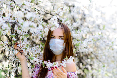Close up portrait of tender girl in a white blouse under a blossoming cherry tree with a mask with...