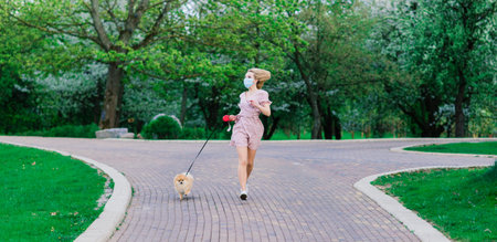 Young woman and red spitz with a medical mask on her face on nature on a spring day. Coronavirus...