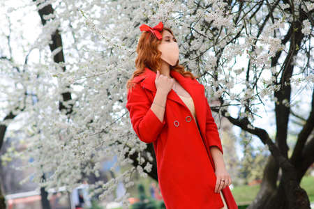 Close up portrait of tender red-haired girl in a red coat under a blossoming cherry tree with a mask from the coronavirus.の写真素材