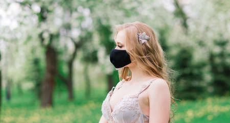 Beautiful young bride in a wedding dress and a black medical mask on her face near a blooming apple trees. Covid-19 protects.の写真素材