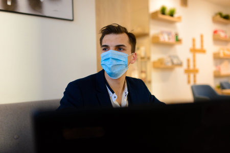 Portrait of young handsome businessman with face mask in quarantine of flu. Photo in caffee with laptop, mobile phone. Coronavirus pandemic.の写真素材