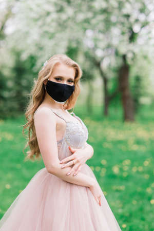 Beautiful young bride in a wedding dress and a black medical mask on her face near a blooming apple trees. Covid-19 protects.の写真素材