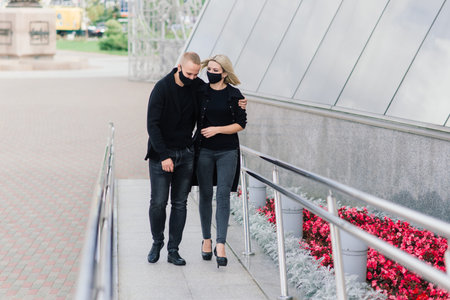 Couple wearing trendy fashionable protective masks, denim jackets, walking in empty street of...