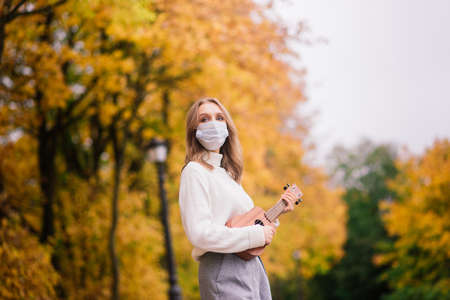Portrait young woman in protective mask playing ukulele guitar in autumn park, lifestyle healthy travel conceptの写真素材