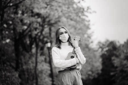 Portrait young woman in protective mask playing ukulele guitar in autumn park, lifestyle healthy travel conceptの写真素材