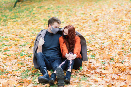 Young red haired woman puts on a face mask while walking with young man in autumn park.の写真素材