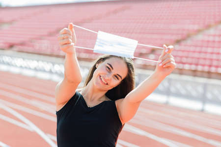 Young fit female in sportswear and protective mask for coronavirus on red track and volleyball ground during outdoor workout at stadiumの写真素材