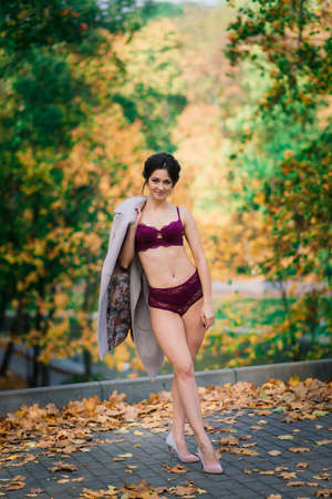 Young beautiful dark-haired woman with a slender figure posing in lingerie and a classic coat in an autumn parkの写真素材