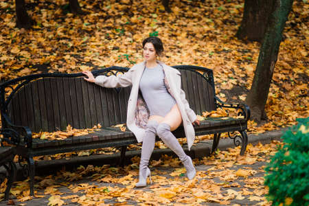 Young beautiful dark-haired woman with a slender figure posing in lingerie and a classic coat in an autumn parkの写真素材