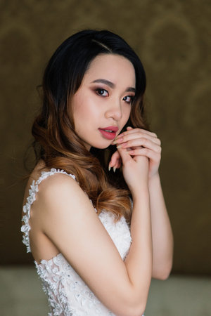 Gorgeous young asian woman in elegant dress looking at camera and window, morning of the bride, weddingの写真素材