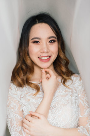 Gorgeous young asian woman in elegant dress looking at camera and window, morning of the bride, weddingの写真素材
