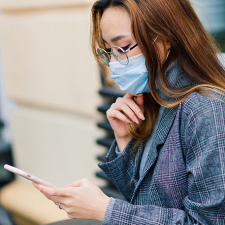 Young Asian businesswoman with mask using phone and sitting with distance outside coffee shopの写真素材