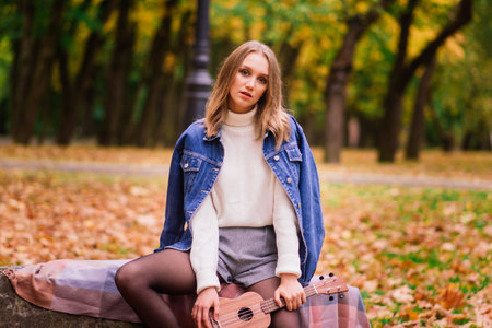 A young woman is sitting on the bench in park and playing ukulele, posing with guitarの写真素材