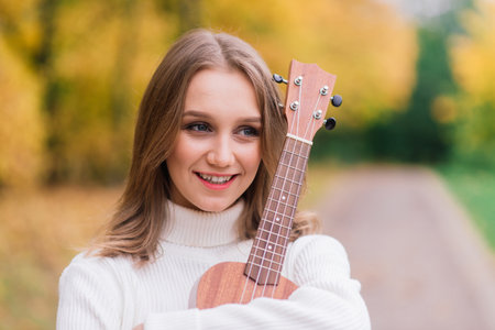 A young woman is sitting on the bench in park and playing ukulele, posing with guitarの写真素材
