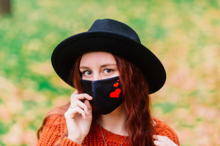 Portrait of young woman in knitted red sweater, hat in a black stylish medical mask in the autumn yellow park. Fashion, lifestyle, quarantine, coronavirus.の写真素材