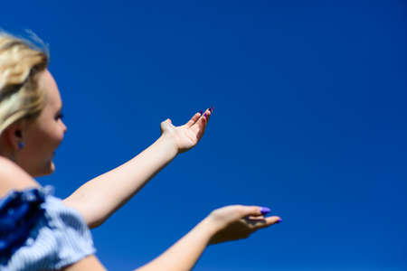 A young blonde girl is looking at the beautiful blue sky and reaches for the sky with her hands. Beautiful female admires nature.