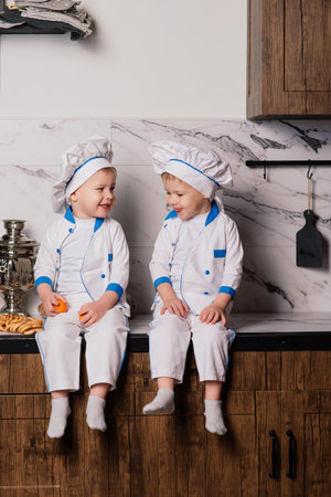 Little cute cook with cutlery sitting on kitchen, twin brothers, tangerine, bagelsの写真素材