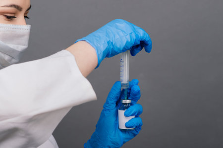 Serious female doctor in a medical mask holding syringe and vaccine isolated on grey