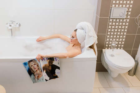 Minsk, Belarus - 01.18.2021. Woman in towel and glasses lying in bath with foam and reads magazineのeditorial素材