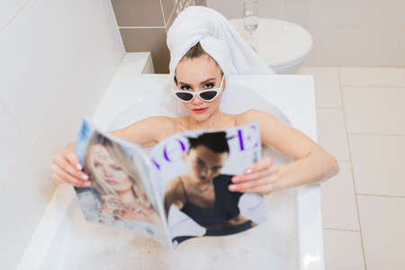 Minsk, Belarus - 01.18.2021. Woman in towel and glasses lying in bath with foam and reads magazineのeditorial素材