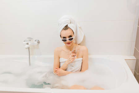 Lovely female with snow-white smile sits in the bathroom. Stylish woman in towel on her head and sunglasses takes spa treatments at home or hotelの写真素材