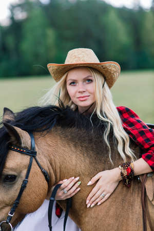 Minsk, Belarus - 09.12.2020. Beautiful blonde young female in elegant hat near the old wooden fence and on field with ponies, country brideのeditorial素材