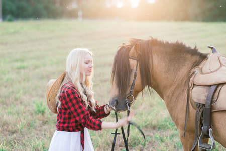 Minsk, Belarus - 09.12.2020. Beautiful blonde young female in elegant hat near the old wooden fence and on field with ponies, country brideのeditorial素材