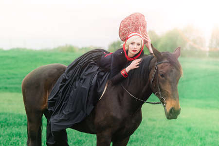 Minsk, Belarus - 05.09.2020. Blonde slavic female in black dress and headdress kokoshnik on field with a black horse at sunsetのeditorial素材