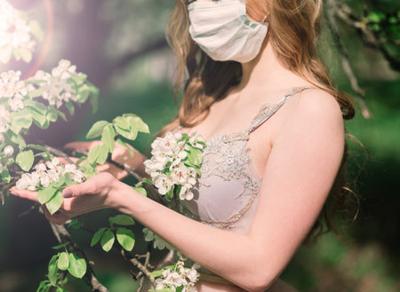 Beautiful young bride in a wedding dress and a white medical mask on her face near a blooming magnolia.の写真素材