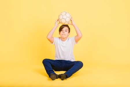 Cute boy is holding a football ball made of genuine leather isolated on yellow background. Soccer