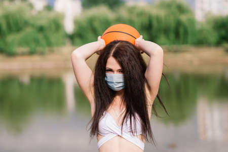 Young sport fitness woman basketball player using mask to protect with ball, lake background.の写真素材