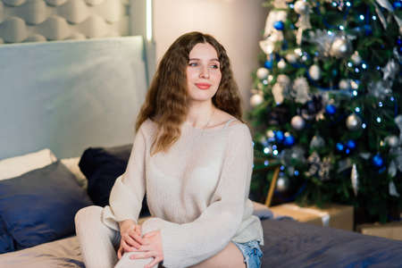 Happy emotional surprised young woman by a Christmas tree in cozy living room, concept of happinessの写真素材