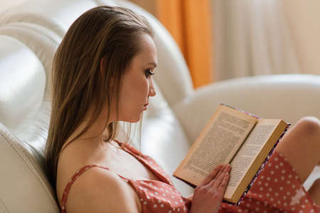 Pensive dreamy female holding book at home looking at big window, thoughtful young woman, studentの写真素材