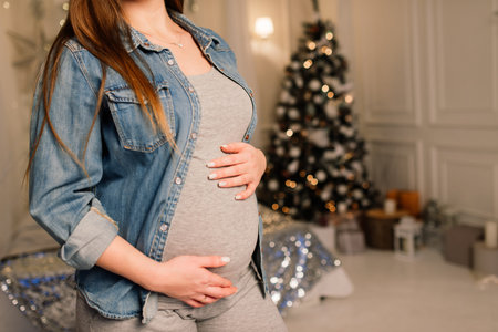 Close up of happy smiling pregnant woman touching her belly over a christmas tree background