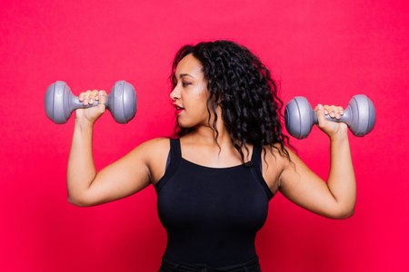 Smiling african american female doing hand exercises with dumbbells on red studio background.の写真素材