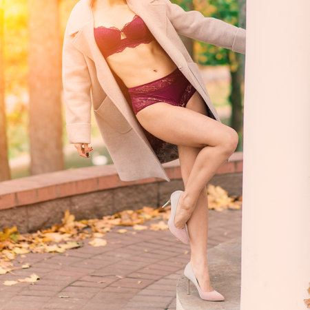 Young beautiful dark-haired woman with slender figure in lingerie and classic coat in autumn parkの写真素材