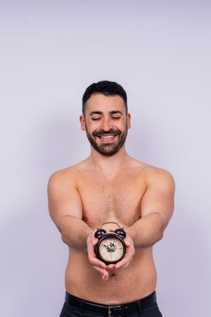Middle-aged white man looks on alarm clock in hand on grey background, copy space for textの写真素材