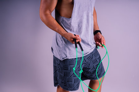 A man with a grey tank top is holding a green jump rope with his elbow bent and waist twisted, showing off his toned chest and wrist strengthの写真素材