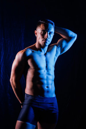A man with a bare chest is posing against a dark backdrop, showing off his muscular physique. His head, hands, stomach, arms, shoulders, legs, and neck are visible in the flash photographyの写真素材