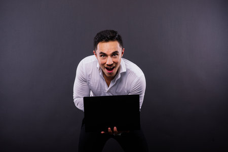 Image of a young cheerful businessman holding and using laptop isolated over dark backgroundの写真素材
