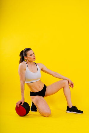 Sportive caucasian cross fit female with fitness ball, look at camera. Studio dark yellow backgroundの写真素材